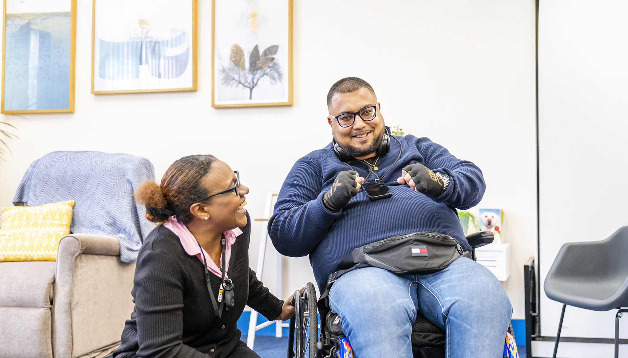 Two people having a funny conversation. A woman is crouched next to the young man in his wheelchair in The Lounge.